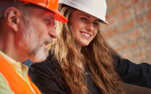 Selected focus photo of young woman in hardhat posing by the side of experienced engineer