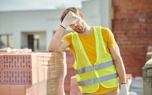 Overwork. Tired man in bright vest taking off his protective helmet and touches face with hand standing near pile of bricks at construction site on fine day